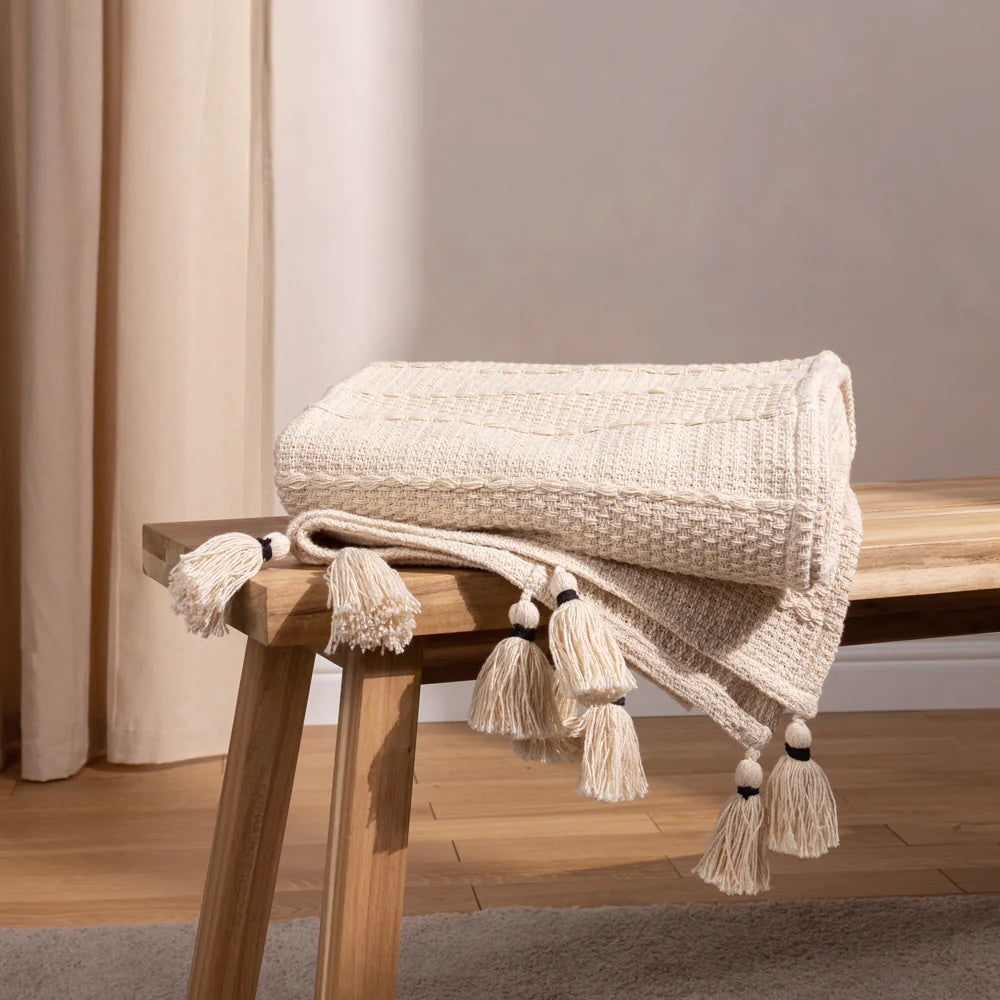 Textured blanket with tassels on a wooden bench against a neutral wall.