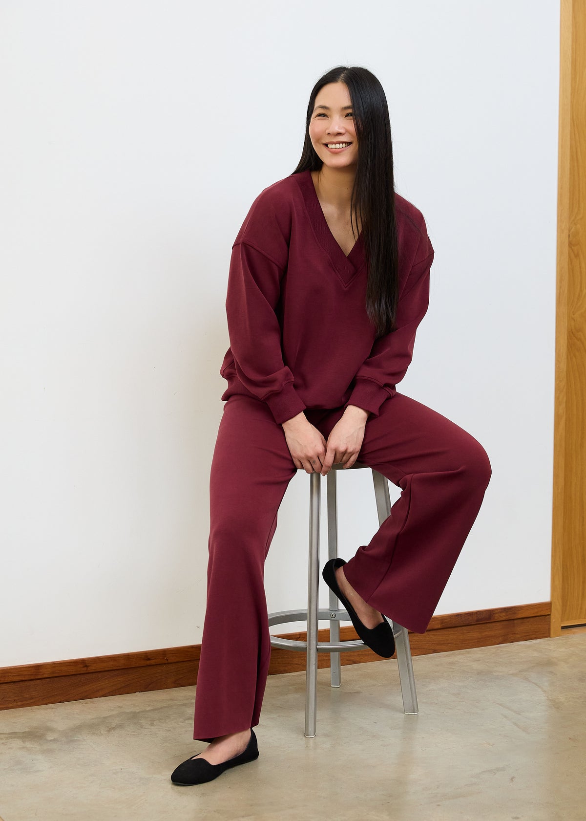 A woman with long black hair sits on a metal stool wearing a matching burgundy lounge set consisting of a V-neck long-sleeve top and wide-leg pants, paired with black flats against a white wall background.